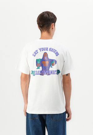 T-shirt blanc en coton avec un graphisme coloré d'une figure de dessin animé tenant des légumes, avec le texte "MANGE TES LÉGUMES VERTS" au-dessus.