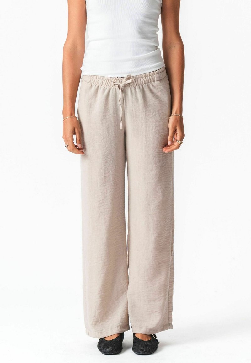Femme portant un pantalon large en lin beige avec une taille à cordon, associé à un haut blanc sans manches et des chaussures noires slip-on.