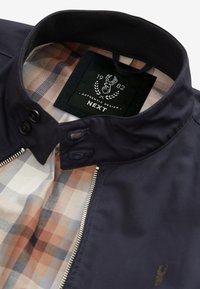 Veste marine avec doublure à carreaux beige, fermeture éclair devant, col boutonné et étiquette noire affichant un emblème de cerf et « 1982 Authentic Design NEXT ».