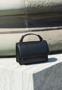 Μαύρη δερμάτινη τσάντα με πάνω λαβή που ακουμπά σε υφασμένη μπεζ πέτρινη επιφάνεια σε εξωτερικό χώρο.