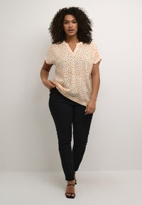 Blouse à manches courtes en orange clair avec des pois disséminés de couleur marron foncé et bleue, accompagnée d'un pantalon noir ajusté et de talons noirs à lanières.