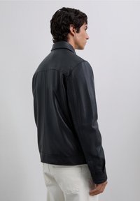 Hombre con chaqueta de cuero negra y pantalones blancos, de perfil frente a un fondo gris claro liso.