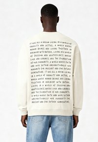 Šviesiai smėlinės spalvos džemperis su ilgomis rankovėmis, ant nugaros turintis drąsų juodą tekstą. Susmulkintas apykaklė ir rankogaliai; laisva forma.