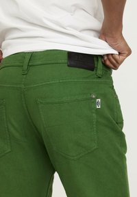 Pantalon en denim vert avec une finition texturée, des coutures visibles et deux poches arrière. Une étiquette en cuir noir est fixée à la taille.