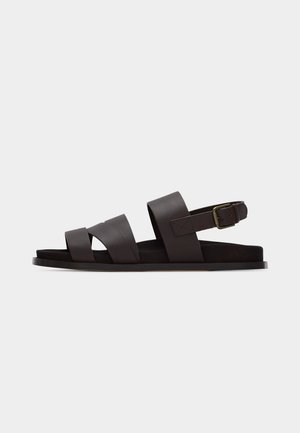 Tmavohnedá kožená sandála s tromi širokými remienkami, nastaviteľným prackovým remienkom okolo členka a plochou podrážkou na bielom pozadí.