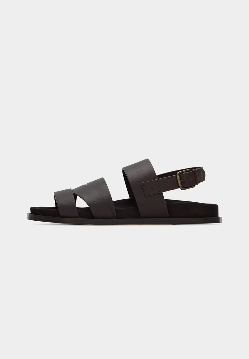 Σκούρο καφέ δερμάτινο σανδάλι με τρεις φαρδιές λουρίδες, ρυθμιζόμενο λουράκι αστραγάλου με αγκράφα και επίπεδη σόλα σε λευκό φόντο.