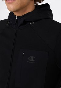 Hombre vistiendo una sudadera con cremallera negra y capucha ajustable, con un bolsillo en el pecho con cremallera que presenta el logo de Champion.
