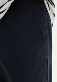 Pantalons bleu marine avec une texture lisse, dotés d'une poche zippée et d'une coupe ajustée, mettant en avant un design minimaliste.