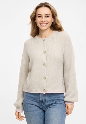 Frau lächelt, trägt eine hellbeige, geknöpfte Strickjacke und blaue Jeans, steht vor einem einfarbigen weißen Hintergrund.