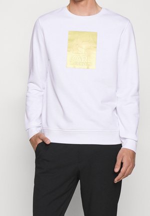 Mężczyzna w białej bluzie z złotym logo Karl Lagerfeld na piersi i czarnych spodniach, jedną ręką w kieszeni.