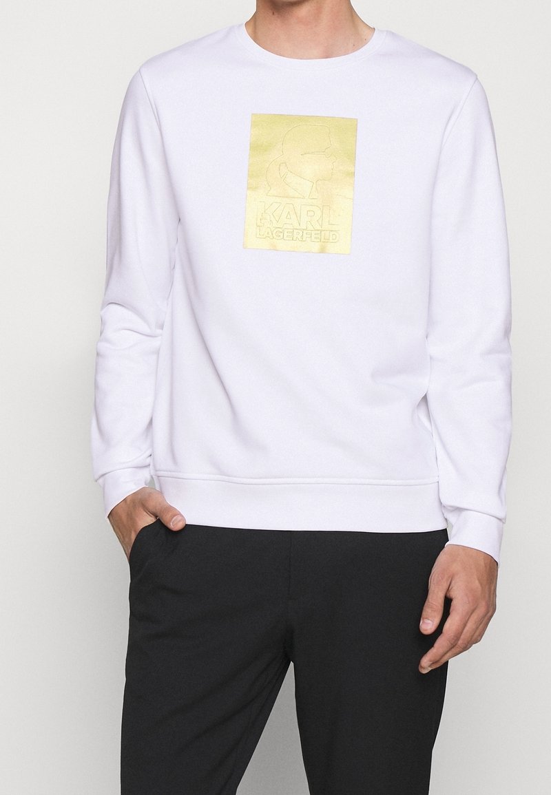 Homme portant un sweat-shirt blanc avec logo doré Karl Lagerfeld sur la poitrine et pantalon noir, une main dans la poche.