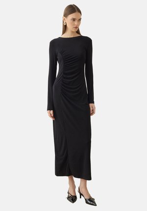 Mujer con un vestido largo negro fruncido con mangas largas y tacones negros de punta, de pie frente a un fondo claro liso.