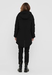 Manteau à capuche noir avec un ourlet arrière légèrement allongé, présentant un détail zippé à l'arrière inférieur, associé à un pantalon noir et des bottes.