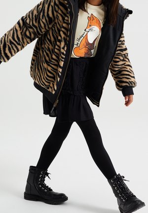 Schwarze Kampfstiefel, ein schwarzer Rock und ein cremefarbener Pullover mit einem Fuchsdesign, kombiniert mit einer beige-schwarzen Jacke mit tigerstreifendem Fellfutter.