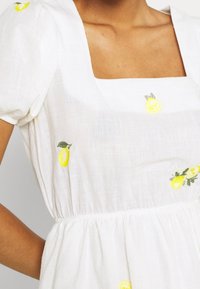 Weißes Kleid mit quadratischem Ausschnitt und Puffärmeln, mit gelben Zitronenstickereien auf dem Stoff. Strukturierter, leichter Stoff.