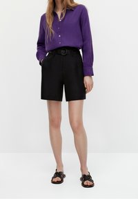Femme portant une chemise boutonnée à manches longues violette, un short taille haute noir avec ceinture et des sandales plates noires, debout les mains dans les poches.