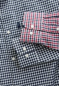 Două cămăși de tip plaid prezintă culori contrastante: albastru și alb cu un fundal bleu marin, roșu și alb cu un fundal burgund, ambele având închidere cu nasturi.