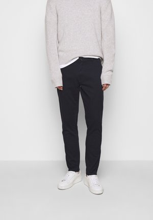 Person, die einen hellgrauen, gestrickten Pullover, schwarze Hose, weißes Unterhemd und weiße Turnschuhe trägt, vor einem schlichten hellen Hintergrund steht.