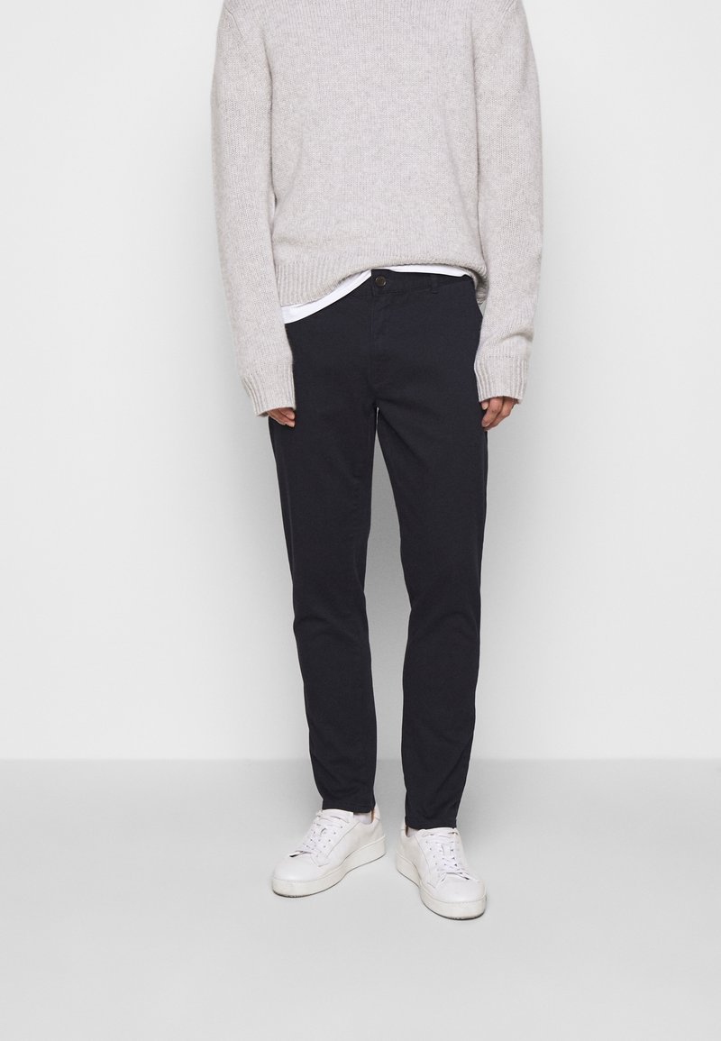 Personne portant un pull en maille gris clair, un pantalon noir, un sous-vêtement blanc et des baskets blanches, debout contre un fond clair uni.