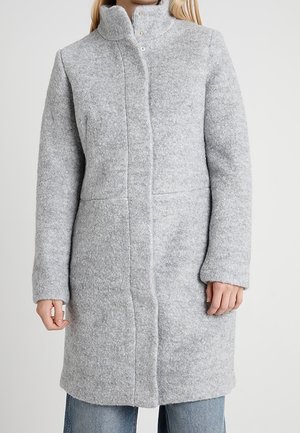 Femme portant un manteau en laine gris clair, longueur genoux, col montant, par-dessus un jean bleu, debout devant un fond clair uni.