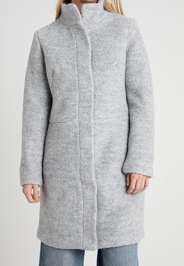 Femme portant un manteau en laine gris clair, longueur genoux, col montant, par-dessus un jean bleu, debout devant un fond clair uni.