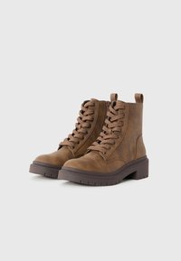 Une paire de bottes chevilles en cuir marron à lacets, avec une fermeture éclair sur le côté et des semelles épaisses et striées de couleur marron foncé, sur un fond blanc uni.