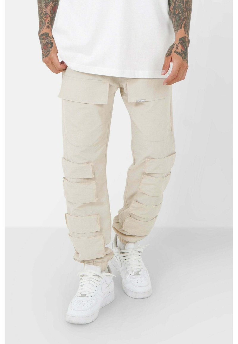 Beige cargobyxor med flera fickor, detaljerad sömnad och smal passform; kombinerat med vita sneakers. Tillverkat i bomull, avslappnad design.
