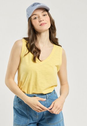 Jeune femme aux longs cheveux bruns portant une casquette de baseball à rayures, un haut jaune sans manches à col en V, et un jean bleu taille haute.