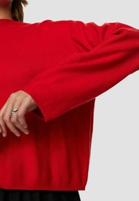 Maglione rosso lavorato a maglia con maniche lunghe, collo rotondo, polsini e orlo a coste. Il tessuto appare morbido con una superficie liscia.
