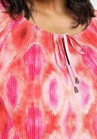 Primo piano di una donna che indossa un top tie-dye rosa e rosso con scollatura arricciata e sottili lacci con estremità argentate.