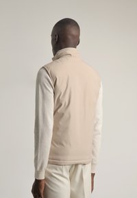 Chaqueta sin mangas color beige superpuesta sobre una camisa de manga larga gris claro. Textura suave, cuello alto y diseño minimalista.