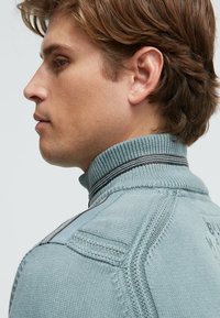 Hellblauer Pullover mit hohem, geripptem Kragen, strukturiertem Strickmuster und schwarzen Streifen am Kragen. Seitenansicht zeigt die Designdetails.