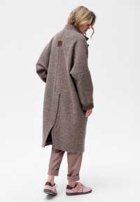 Bruine houndstooth jas met een relaxte pasvorm, voorzien van een hoge kraag, zijsplitsingen en een bruin leren merklabel op de achterkant. Gecombineerd met beige broek en roze sneakers.