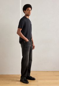 Schwarzes kurzärmeliges T-Shirt kombiniert mit dunkelgrauen geraden Jeans. Das Modell steht auf einem Holzboden mit den Händen in den Taschen und trägt schwarze Schuhe.