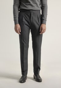 Hombre vestido con pantalones plisados gris oscuro, suéter gris y zapatos de vestir negros, de pie contra un fondo liso claro.