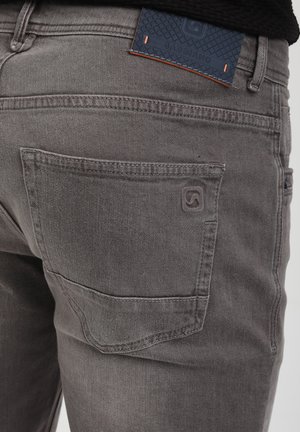 Gray denim jeans with a textured blue leather tag on the waistband, featuring a back pocket with a subtle logo detail.
