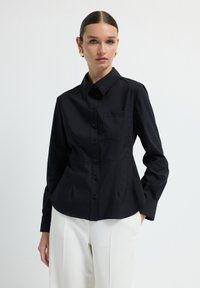 Una donna con capelli lisci indossa orecchini d'oro, una camicia nera aderente con bottoni e pantaloni bianchi, in piedi contro uno sfondo chiaro e semplice.