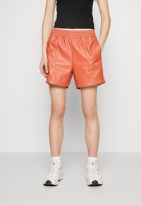 Orange faux leather shorts with an elastic waistband, side pockets, and snap closures at the hem. Accompanied by white athletic shoes.