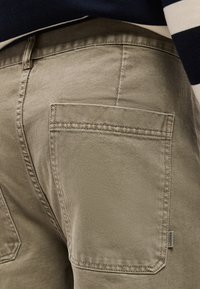 Κοντινό πλάνο τσέπης μπεζ παντελονιού με ορατή ραφή και μικρή ετικέτα υφάσματος μάρκας στο πλάι, φορεμένο με σκούρο ριγέ τοπ.