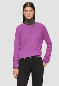 Lila strukturiertes Sweatshirt mit langen Ärmeln, rundem Ausschnitt, über einem dunkelgrauen Rollkragenpullover getragen, kombiniert mit schwarzen Jeans.