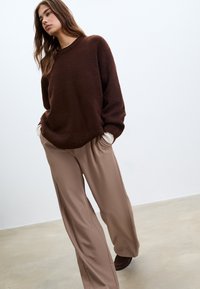 Brauner, strukturierter Pullover mit Rundhalsausschnitt, kombiniert mit weiten beigen Hosen. Das Modell posiert mit den Händen in den Taschen vor einem neutralen Hintergrund.