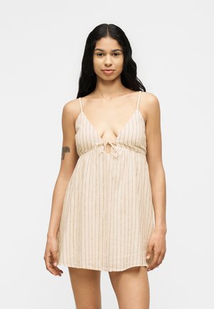 Femeie cu păr lung și închis la culoare, purtând o rochie bej fără mâneci, cu dungi verticale subțiri roșii și un detaliu cu nod în față, stând pe un fundal simplu.