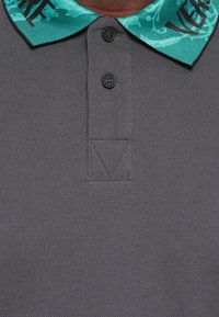 Grå polo-skjorte med ett mønstret teal krage. Har to svarte knapper og en trekantet sømdetalj nederst. Tekst på kragen er synlig.