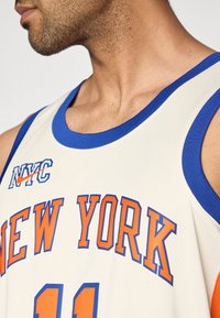 Weißes Basketballtrikot mit blauem Rand, darauf in orangefarbener Schrift "NEW YORK" und "NYC". Unten auffällige Nummer "11". Netzstruktur.