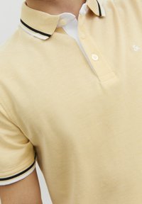 Keltainen polo-paita, jossa on tekstuuri, valkoinen kaulus ja mustat raidat kauluksessa. Varustettu kahdella napilla.