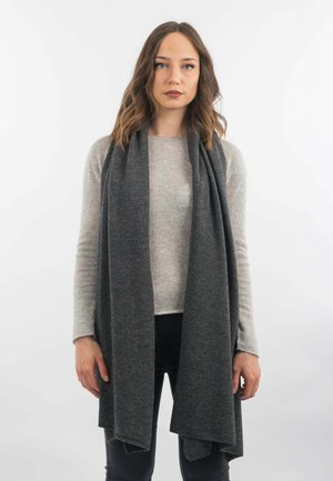 Sciarpa di lana grigia drappeggiata sulle spalle, con texture morbida e forma rettangolare lunga. Il modello indossa un maglione chiaro e pantaloni scuri su uno sfondo bianco.