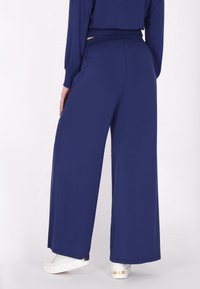 Wijduitlopende broek in navyblauwe stof met een gladde textuur en een elastische tailleband. Gecombineerd met witte sneakers met gouden accenten.