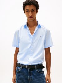Jovem vestido com uma camisa de botão de manga curta azul clara sobre uma camiseta branca, combinada com jeans azul escuro e um cinto trançado preto.