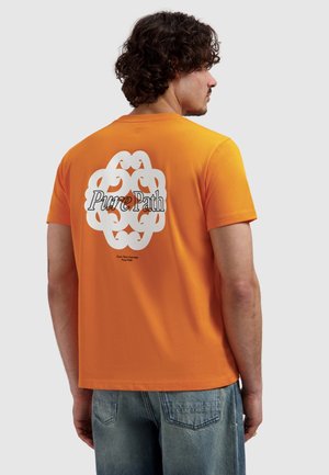 Man met krullend haar die een oranje T-shirt draagt met een wit grafisch ontwerp en de tekst "Pure Path" op de rug, gecombineerd met lichtblauwe spijkerbroek.