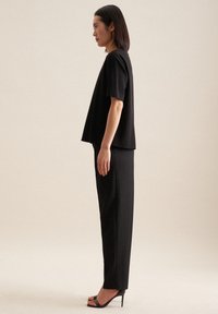 Top nero dalla vestibilità rilassata con maniche corte abbinato a pantaloni neri su misura. Modella che indossa tacchi neri aperti. Sfondo neutro.